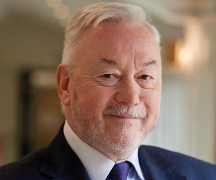 A senior person with short white hair and a beard is wearing a dark suit and tie while smiling softly in a softly lit indoor setting. Description generated by AI