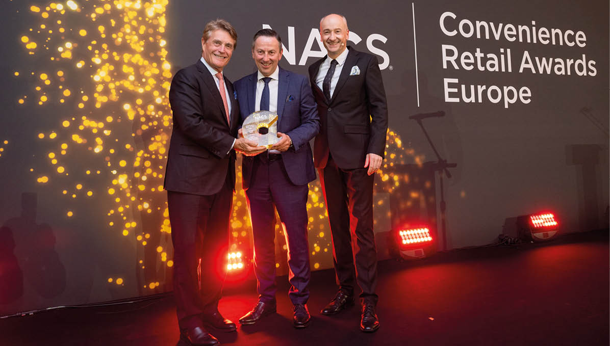 Three adults in formal suits stand on a stage holding an award with a backdrop displaying “NACS Convenience Retail Awards Europe" and decorative golden lights. Description generated by AI