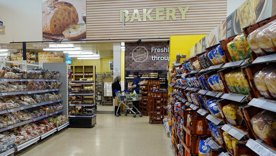 A grocery store aisle with shelves stocked with various packaged bread products and people shopping in the background. Description generated by AI