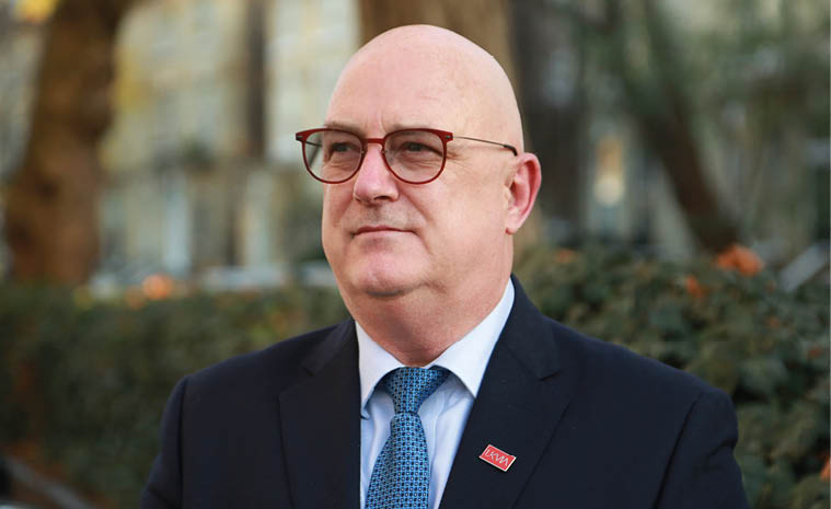 A person wearing glasses, a suit, and a blue patterned tie is standing outdoors with trees and buildings blurred in the background. Description generated by AI