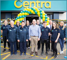 A group of people wearing casual and uniform clothing stand smiling in front of a store entrance decorated with a yellow and green balloon arch and surrounded by racks of flowers. Description generated by AI