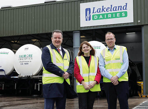 Three people wearing high-visibility vests stand smiling in front of a Lakeland Dairies building with tanker trucks parked nearby. Description generated by AI