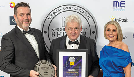 Three people are standing in front of a large poster. A man is holding a framed award, while a woman stands beside him. The woman is wearing a blue dress. AI generated content