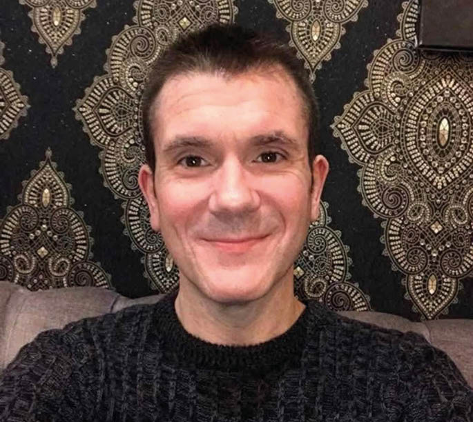 A man is sitting on a couch with a black and white patterned wall behind him. He is smiling and looking at the camera. AI generated content