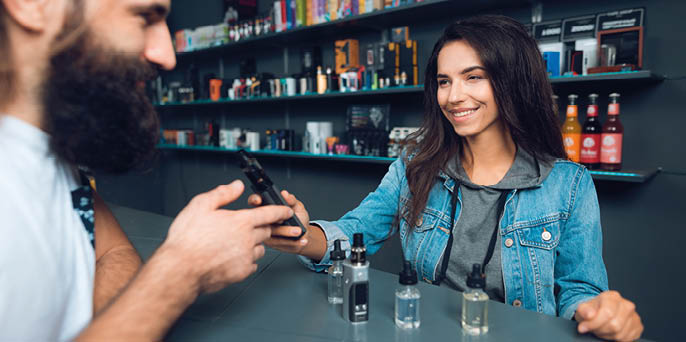 Girl seller shows the choice of electronic cigarettes in vapeshop. Nearby is a buyer - a man with a beard. The store has a large assortment of electronic cigarettes.