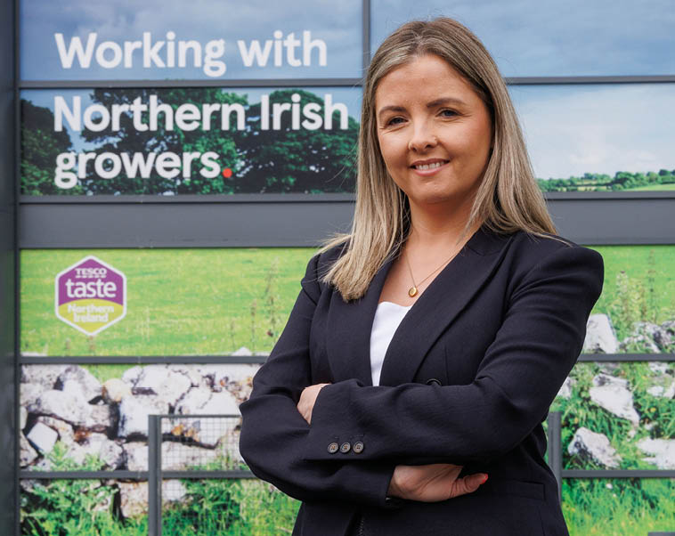 A woman in a black suit standing in front of a sign for Northern Irish growers. Description generated by AI