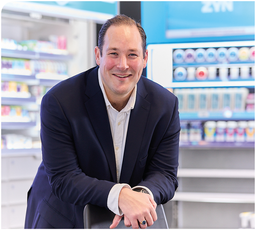 A man wearing a blue jacket and white shirt is smiling in front of a pharmacy. Description generated by AI