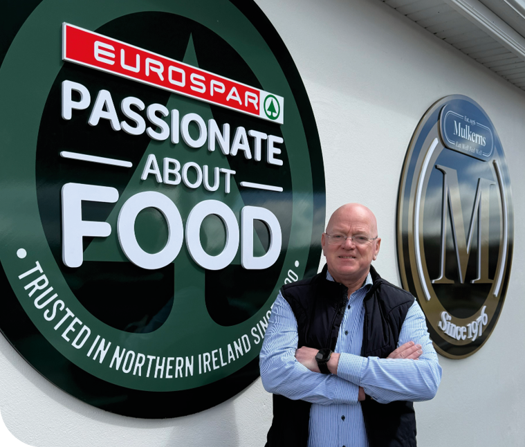 A man standing in front of a green sign that says “Eat Passionate About Food". Description generated by AI