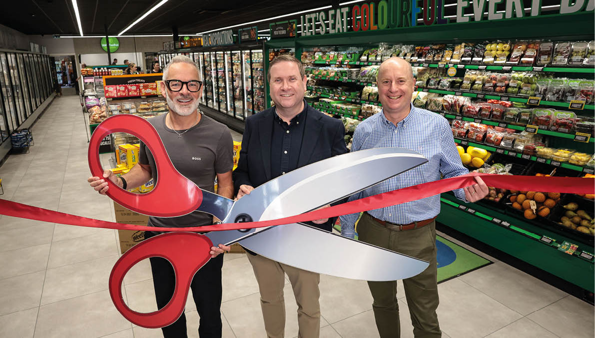 Three adults smiling and holding a large pair of scissors cutting a red ribbon inside a grocery store. Description generated by AI