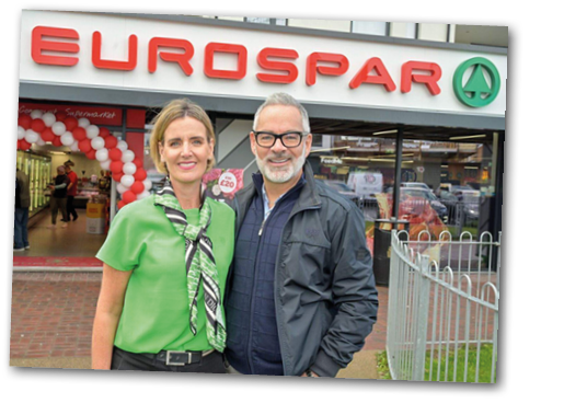 Two adults smiling and standing close together outside a Eurospar supermarket with balloons visible inside the entrance. Description generated by AI