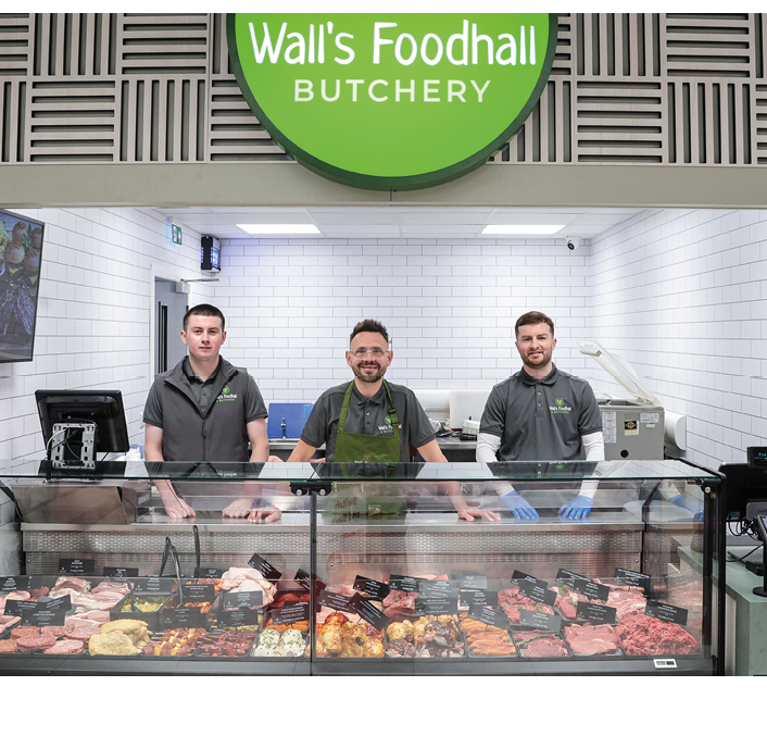 Three adults wearing gray shirts stand behind a glass display case filled with various cuts of meat inside a butcher shop with a green sign reading Wall's Foodhall Butchery above them. Description generated by AI