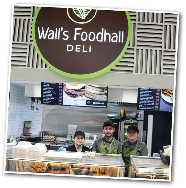 Three people wearing green aprons and black caps stand behind the counter of a deli named Wall's Foodhall with food displays and menu boards visible in the background. Description generated by AI