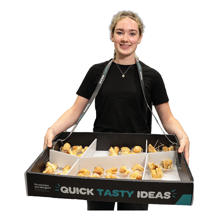 A person wearing a black shirt is smiling and holding a tray with multiple compartments filled with small baked snacks labeled “Quick Tasty Ideas." Description generated by AI