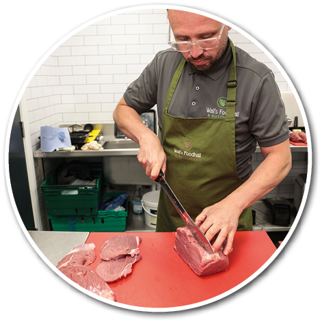 A person wearing glasses and an apron is cutting a large piece of raw meat on a red cutting board in a kitchen setting. Description generated by AI