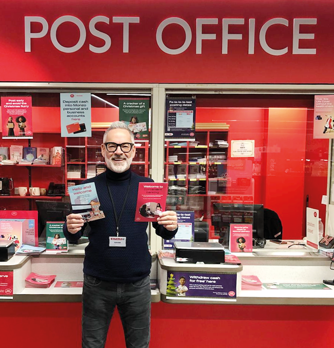 A person wearing glasses and a dark sweater stands behind a counter in a post office holding two informational pamphlets with a bright red and white post office sign above. Description generated by AI
