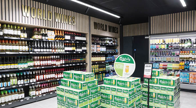 Shelves stocked with various bottles of wine and beverages surround stacks of green Carlsberg beer boxes in a well-lit store aisle labeled World Wines. Description generated by AI