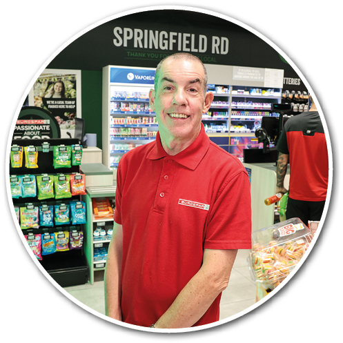 A person wearing a red polo shirt stands smiling inside a store with shelves of products and a sign that reads Springfield Rd in the background. Description generated by AI
