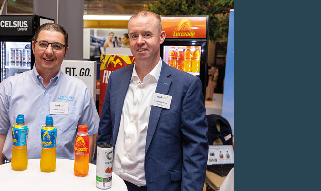 Two adults wearing name tags stand behind a table with bottles of orange and yellow beverages in a setting that appears to be a promotional or trade event. Description generated by AI