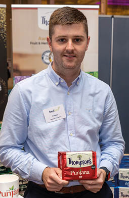 A person wearing a light blue button-up shirt is holding a red package of Thompsons Punjana tea while standing indoors near a display of similar products. Description generated by AI