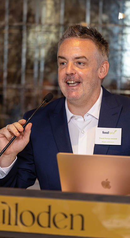 A person wearing a dark blue suit and white shirt is speaking into a microphone while standing behind a podium with a laptop and a name tag that reads “food Paris Island." Description generated by AI