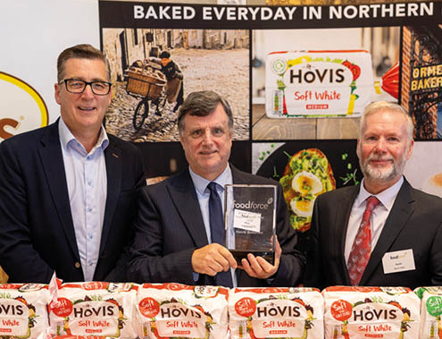 Three adults in business attire stand behind a table displaying multiple loaves of Hovis Soft White bread with a promotional backdrop featuring bread and baking images. Description generated by AI
