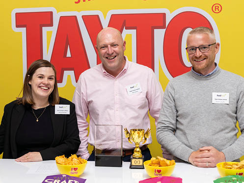 Three adults smiling and sitting behind a table with bowls of snacks and trophies in front of a bright yellow background with large red text. Description generated by AI