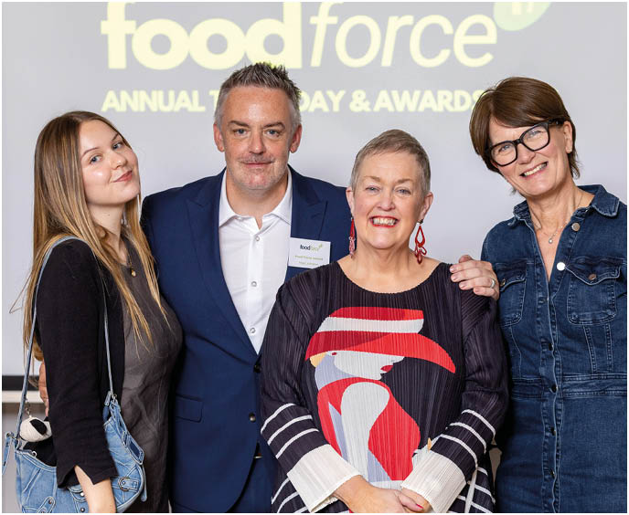 Four adults stand closely together smiling in front of a screen displaying the words food force annual today and awards, with one person wearing a colorful patterned top and another in a denim dress. Description generated by AI