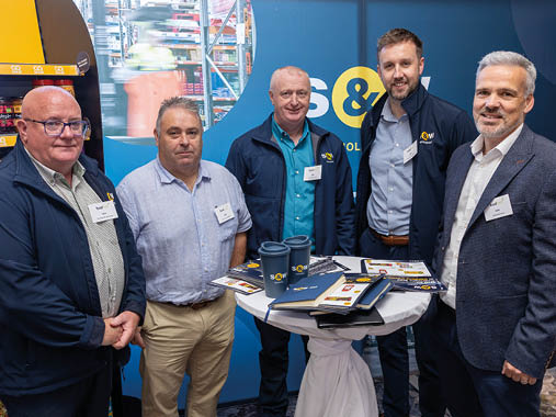 Five adults wearing business casual clothing stand around a tall round table with promotional materials and branded cups in a professional event setting. Description generated by AI