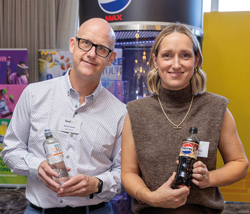 Two adults smiling and holding bottles of beverages while standing indoors in front of promotional displays. Description generated by AI