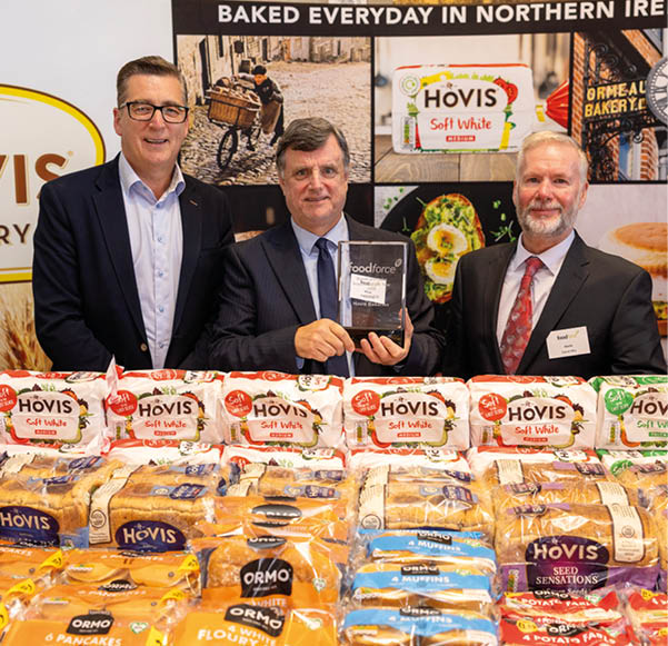 Three adults in suits stand behind a table filled with various packaged Hovis bread products, with one person holding an award plaque in front of a promotional backdrop. Description generated by AI