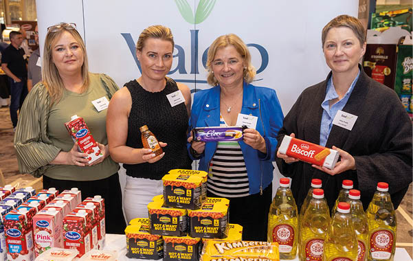 Four adults stand behind a table displaying various packaged food products while holding some of the items and smiling in a well-lit indoor setting. Description generated by AI