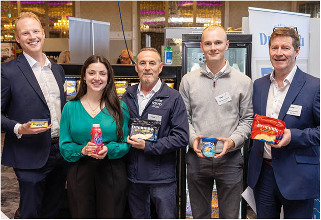 Five adults standing indoors smiling and holding various packaged food and drink products in a professional or promotional setting. Description generated by AI