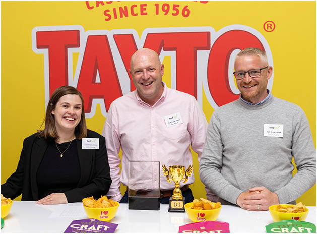 Three adults smiling and standing behind a table with various colorful bags of Tayto snacks and bowls of chips in front of a yellow wall with a large Tayto logo and the text “Castle Cooked Since 1956." Description generated by AI
