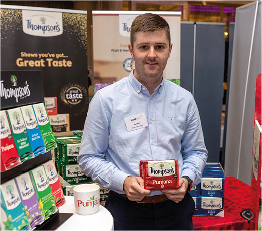 A person wearing a light blue button-up shirt stands behind a table displaying various colorful packaged products from Thompson's, holding one package labeled “Punjab." Description generated by AI