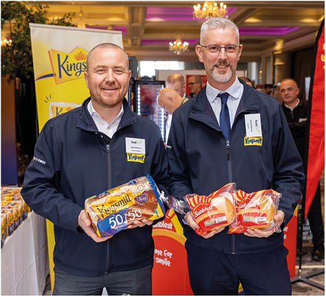 Two adults wearing navy jackets stand indoors holding packaged bread products with promotional banners in the background. Description generated by AI