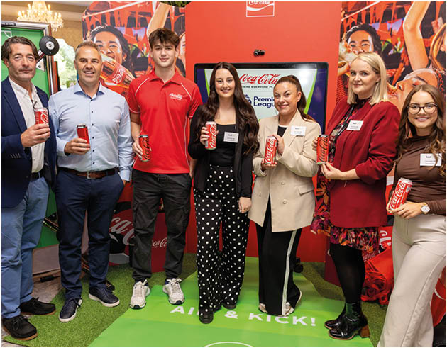 Seven people standing indoors on artificial grass in front of a Coca-Cola branded backdrop, each holding a can of Coca-Cola and smiling. Description generated by AI