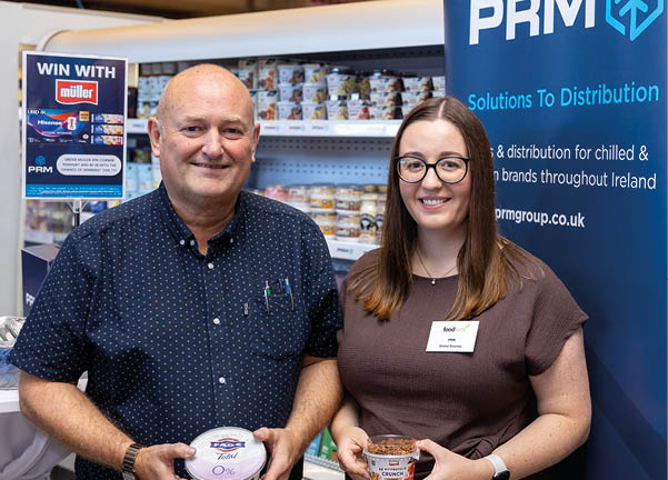 Two adults stand smiling in front of a display of packaged food products and a blue banner with the text PRM Solutions To Distribution, each holding a small food container. Description generated by AI