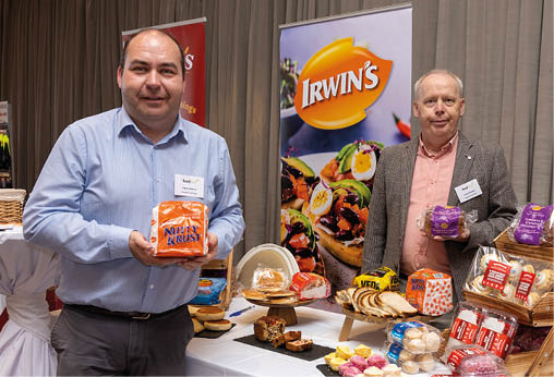 Two adults stand behind a table displaying various packaged baked goods and desserts at a promotional event with branded banners in the background. Description generated by AI