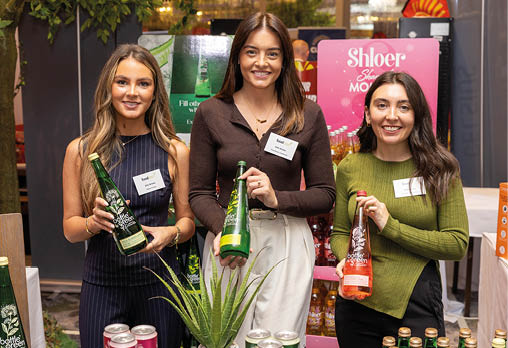 Three adults standing behind a table displaying various bottles and cans of beverages, smiling and holding bottles, with a plant in the center and promotional signs in the background. Description generated by AI