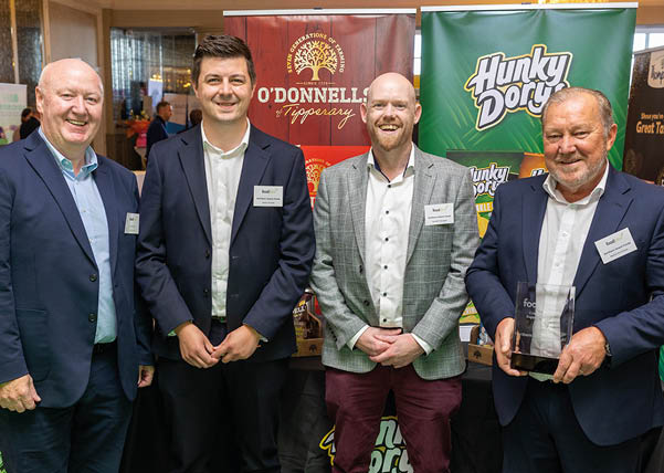 Four adults dressed in business attire stand smiling in front of promotional banners, with one holding a clear award. Description generated by AI