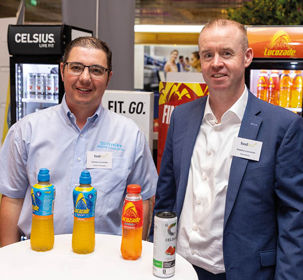 Two adults wearing name tags stand behind a table displaying bottles of sports drinks in a promotional setting with branded refrigerators in the background. Description generated by AI
