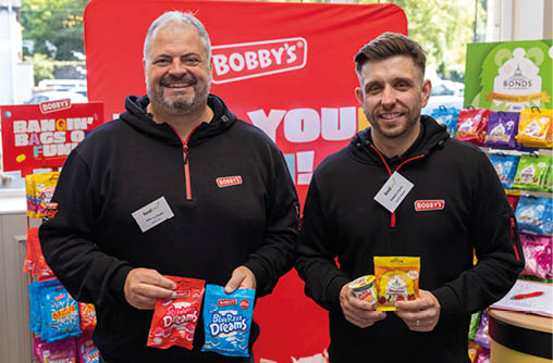 Two adults wearing black hoodies with name tags smile while holding colorful snack bags in front of a display of various packaged snacks and a red backdrop with the word Bobby's. Description generated by AI