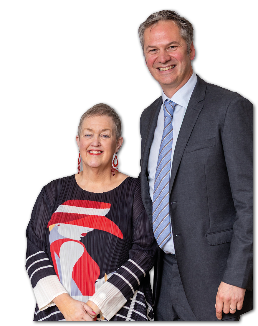 Two adults are standing close together smiling, one wearing a dark suit with a striped tie and the other wearing a colorful patterned dress with red earrings. Description generated by AI