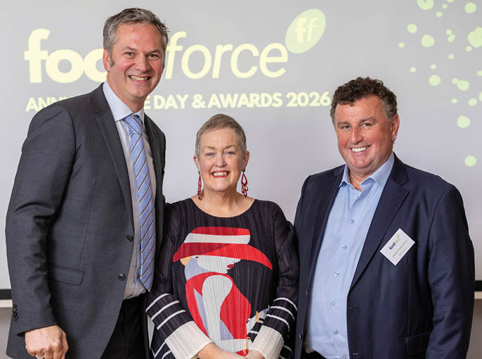 Three adults dressed in business attire stand smiling in front of a backdrop displaying text about an awards event. Description generated by AI