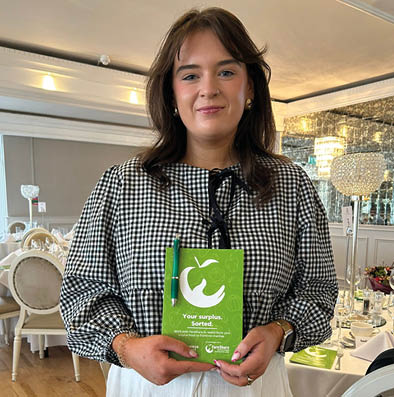 A person with shoulder-length hair wearing a black and white checkered blouse and white pants is standing indoors holding a green booklet with a white apple logo and text. Description generated by AI