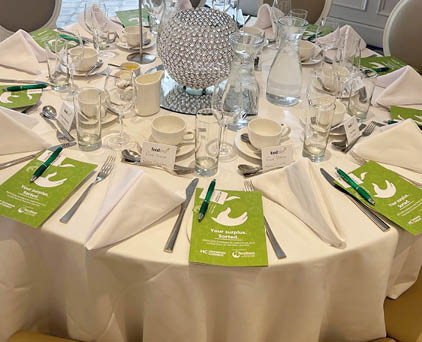 A round table set for a formal event with neatly folded white napkins, green informational cards, glassware, cutlery, and a decorative centerpiece made of a spherical arrangement of crystals. Description generated by AI