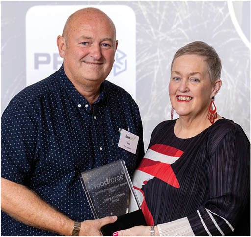 Two adults are smiling and holding a clear award plaque together in a formal setting with a presentation screen in the background. Description generated by AI