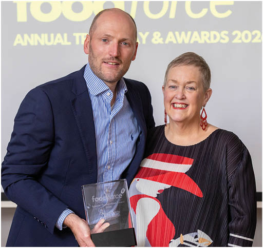 Two adults stand side by side smiling, with one holding a clear award plaque in front of a screen displaying “food force ANNUAL TIMELINE & AWARDS 2026." Description generated by AI