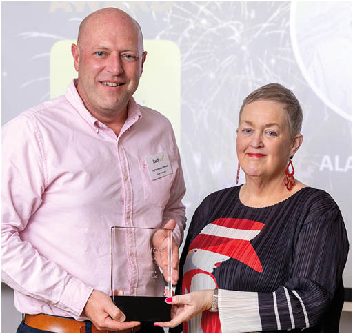 Two adults stand side by side indoors, smiling and holding a clear rectangular award together, with a presentation screen in the background. Description generated by AI