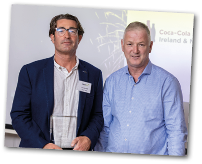 Two adults dressed in business casual attire stand side by side, with one holding a clear rectangular award in front of a presentation screen. Description generated by AI